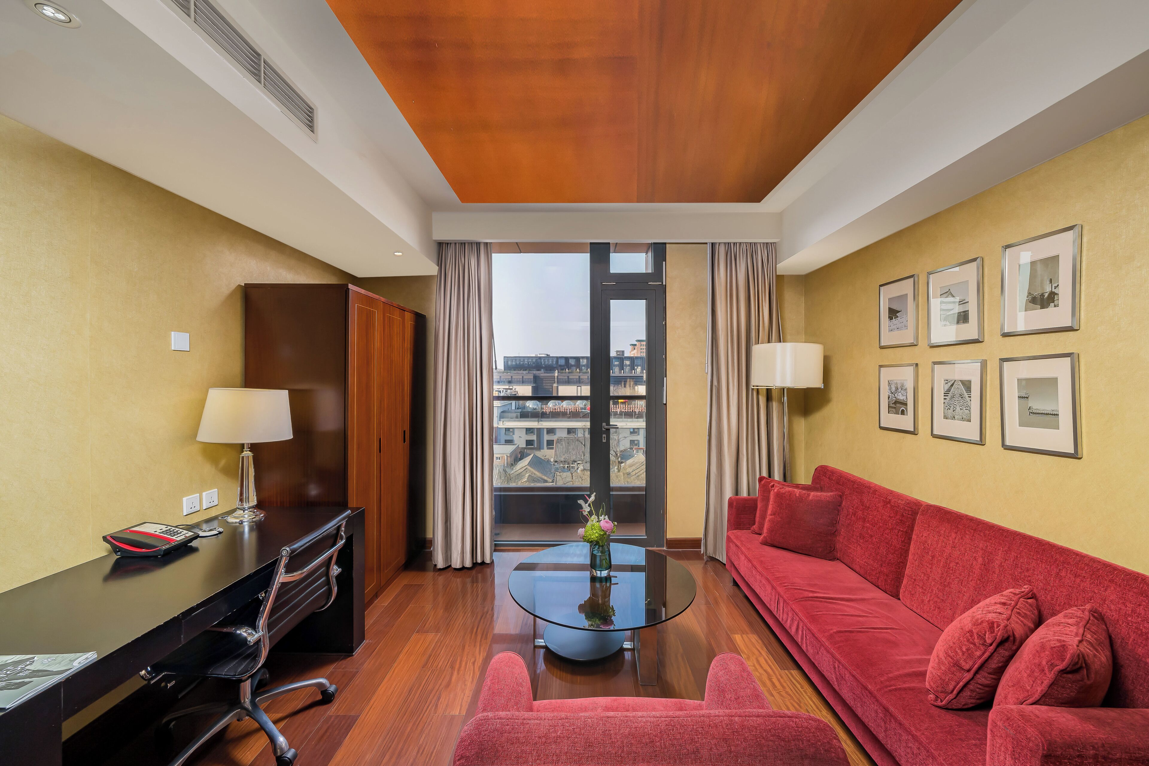 Une chambre d’hôtel équipée d’un bureau, d’un canapé rouge, d’une table basse en verre et offrant une vue sur la ville grâce à de grandes fenêtres.