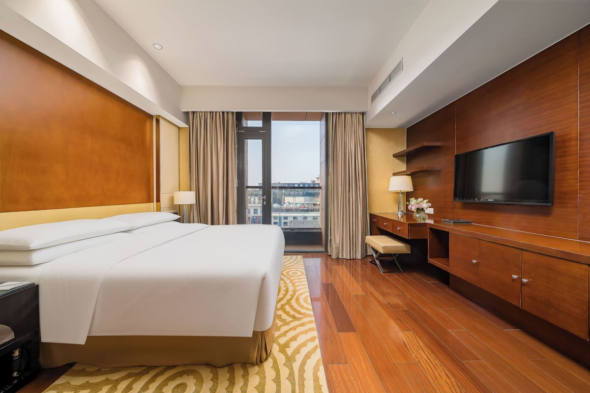 A hotel room with a bed, a TV, a desk, and a view of the city through large windows.