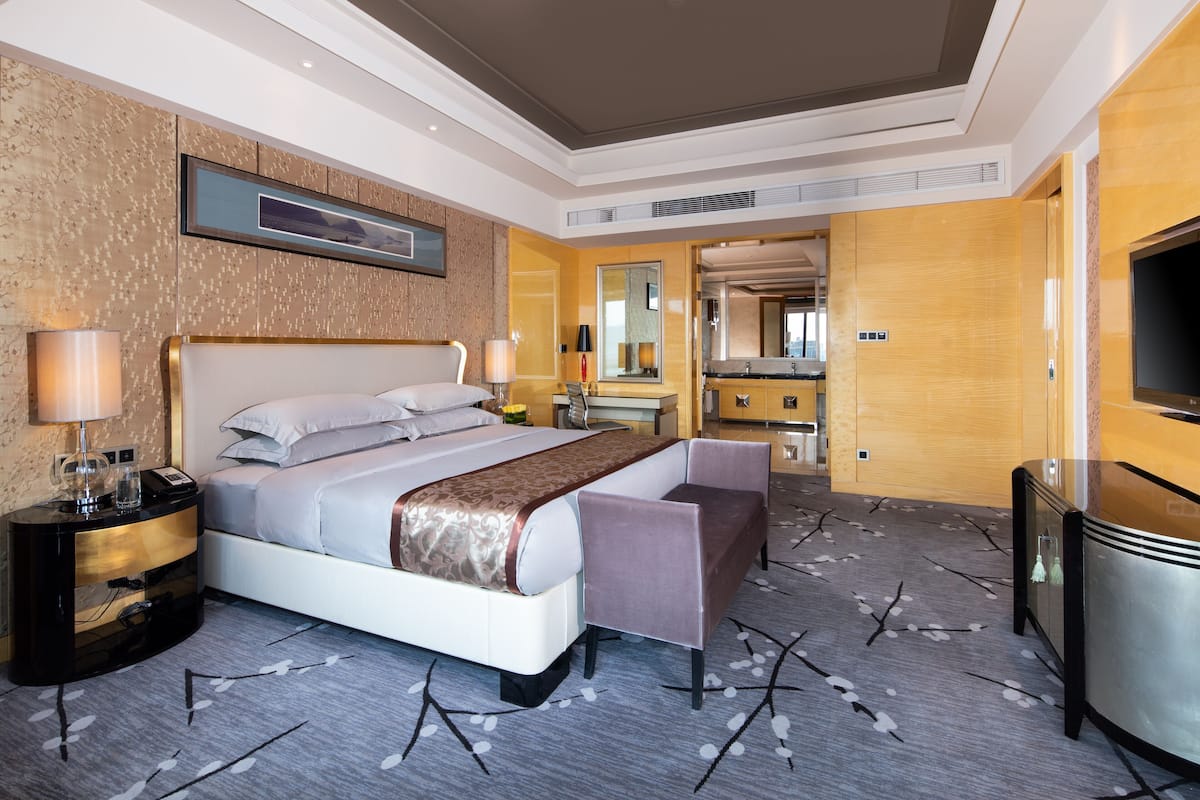 Une chambre d’hôtel avec un grand lit, des tables de chevet, un bureau, une chaise, une télévision et une salle de bain visible à travers une porte ouverte.