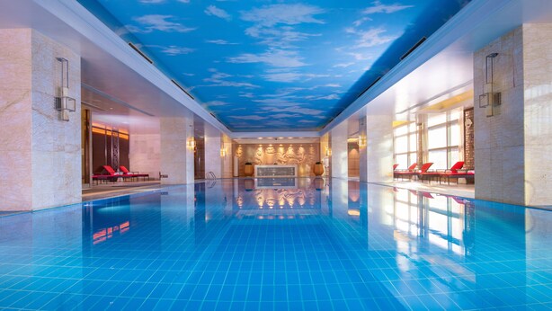 A spacious indoor swimming pool with a clear blue floor, red lounge chairs, and a ceiling with a cloud design. - Wyndham Grand Plaza Royale Colorful Yunnan Kunming (Kunming)