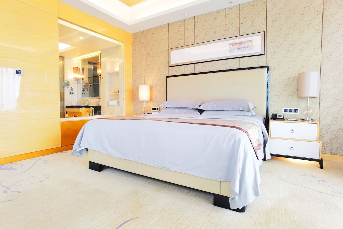 Une chambre d’hôtel moderne avec un grand lit, une table de chevet, une commode et une salle de bain visible à travers une porte ouverte.