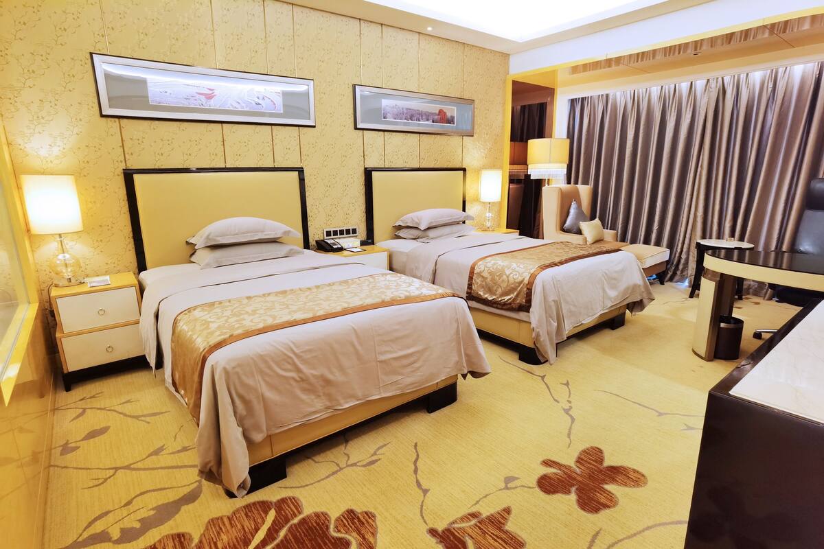 Une chambre d’hôtel avec deux lits, un bureau, une chaise, une lampe et une grande fenêtre avec des rideaux.