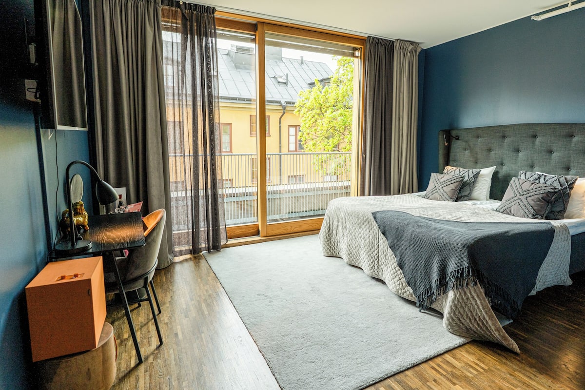A bedroom with a large bed, a desk, and a chair. There is a window with curtains and a view of buildings outside.