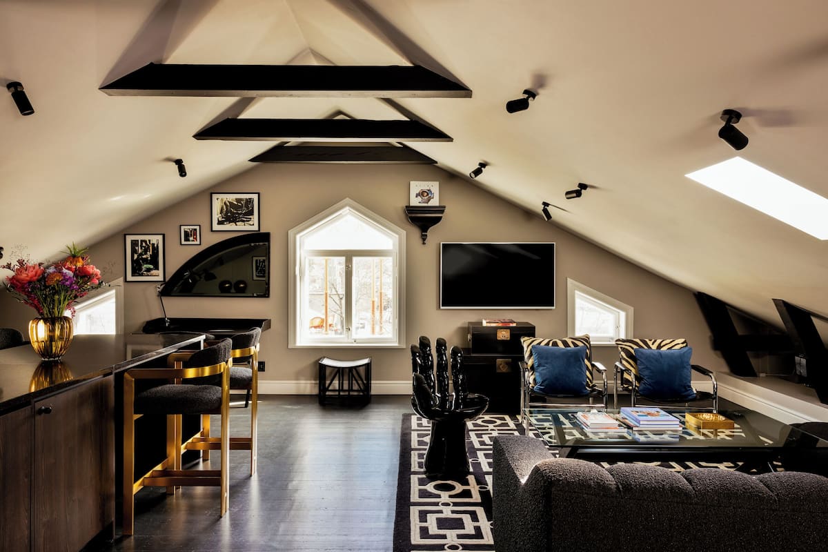 An attic living space with a kitchen area, a seating area with a sofa and chairs, a dining table with books, and a large window providing natural light.
