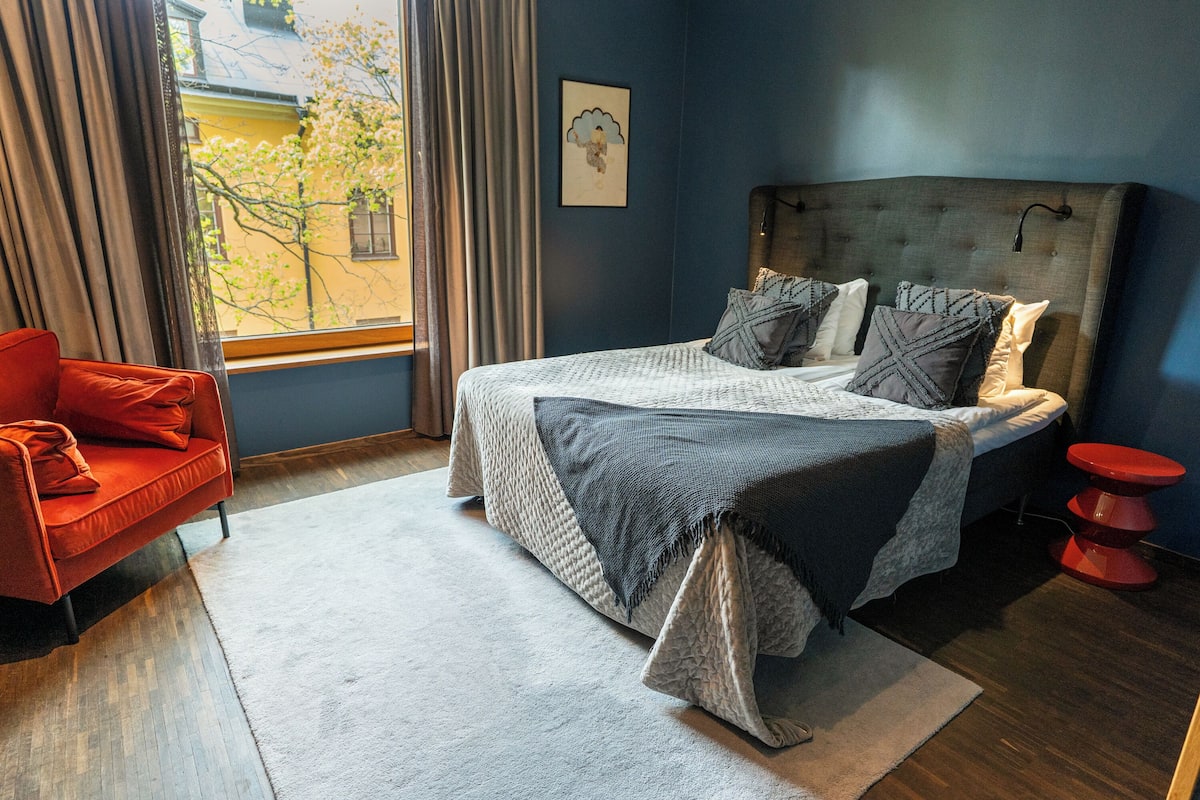 A bedroom with a bed, a red chair, a window with curtains, and a view of a building outside.