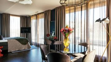 Habitació d'hotel moderna amb llit, taula de dinar amb caires, televisió i una finestra gran amb cortines.