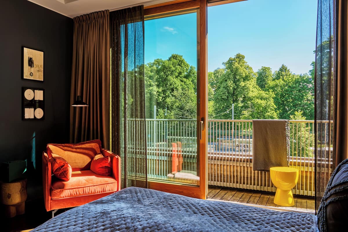 A bedroom with a large bed, a red sofa, a yellow stool, and a balcony with a view of trees.