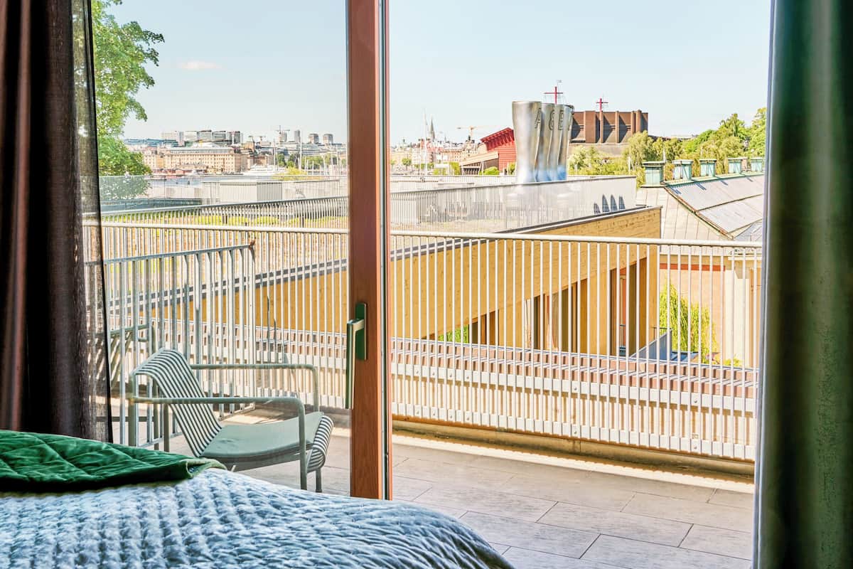 A balcony with a metal railing, a bed with a green blanket, and a view of a cityscape with buildings and a bridge.