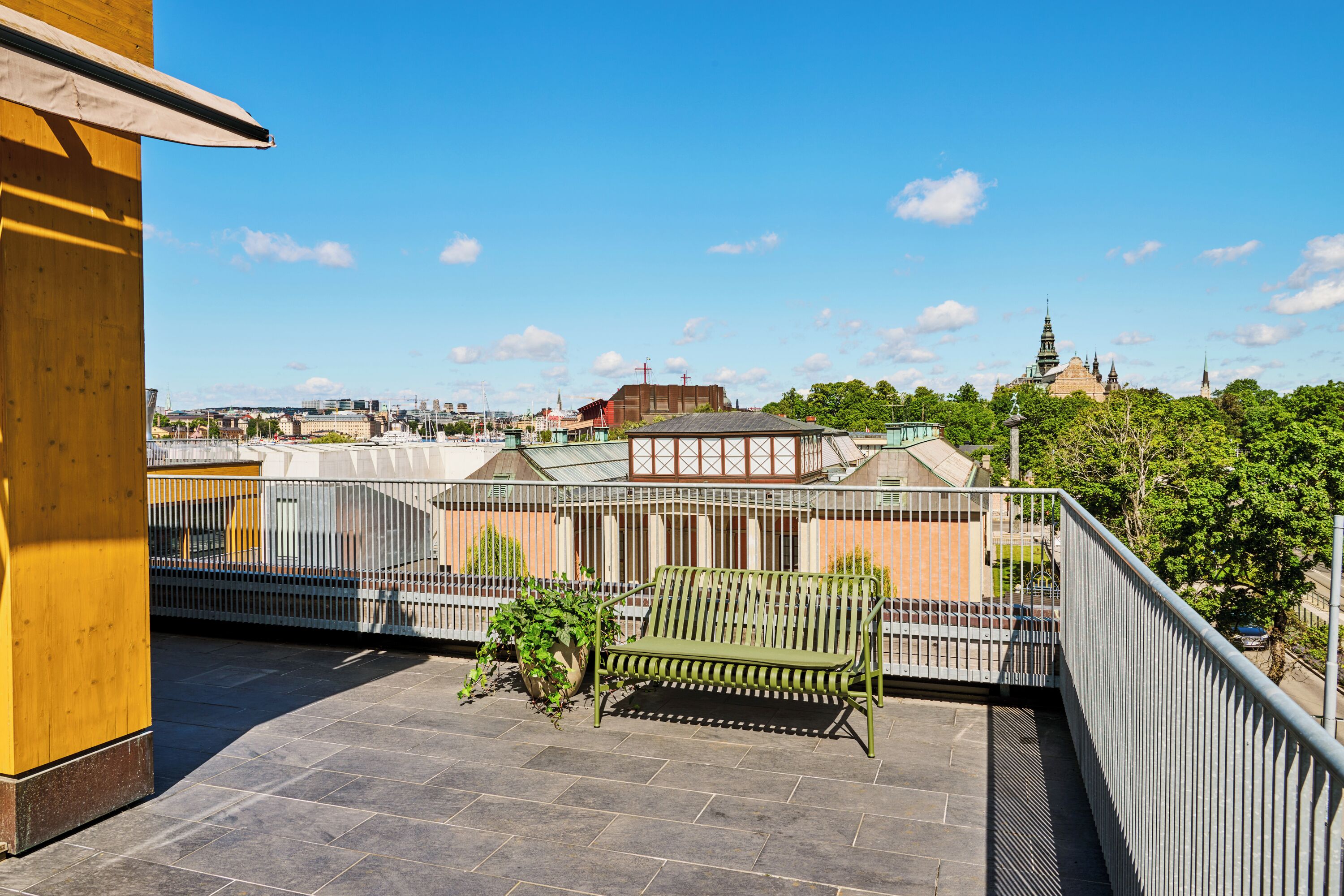 A balcony with a green bench, a potted plant, and a view of a cityscape with buildings and a church.