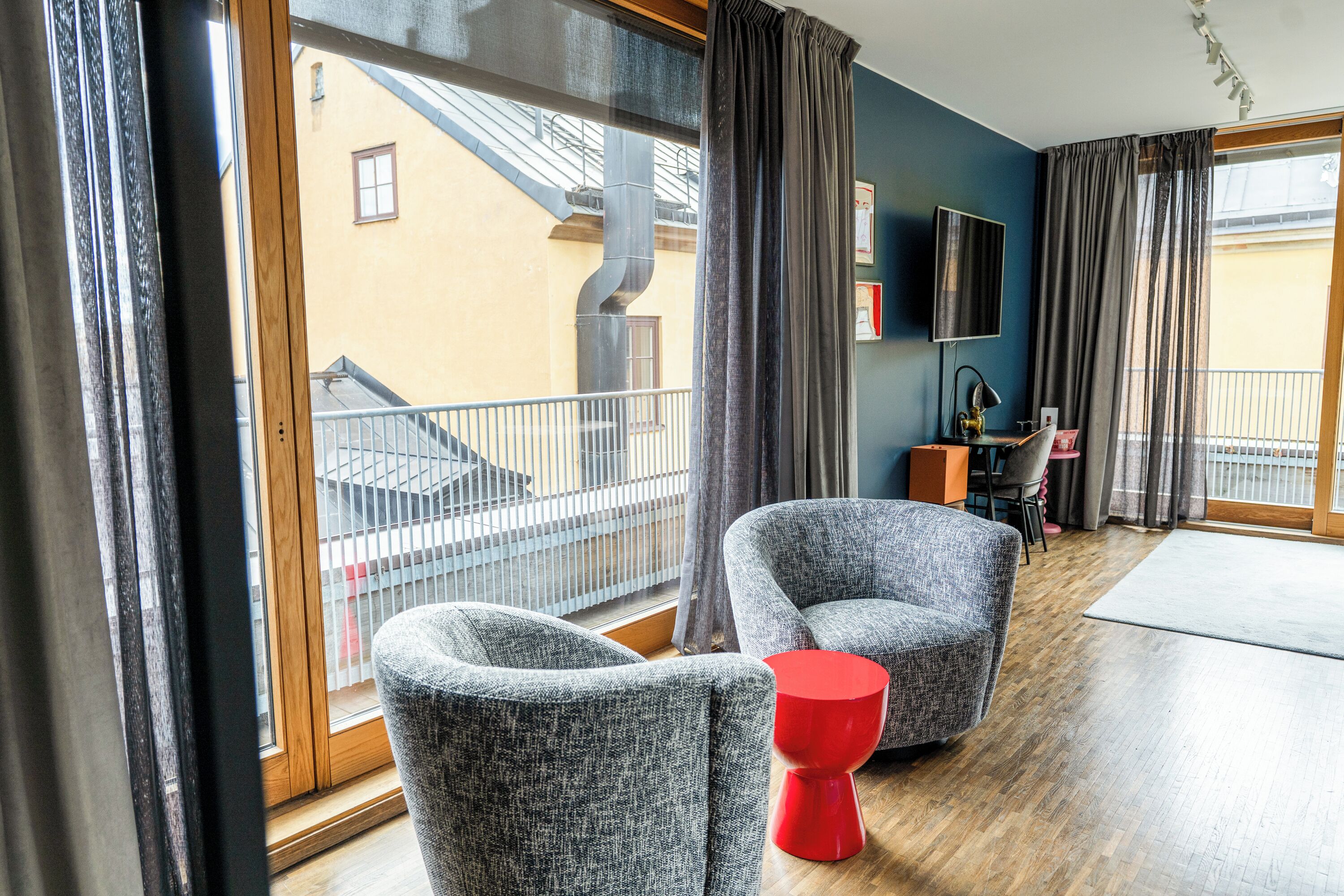 Ein modernes Hotelzimmer mit großen Fenstern, zwei grauen Sesseln, einem roten Beistelltisch und einem Holzboden.
