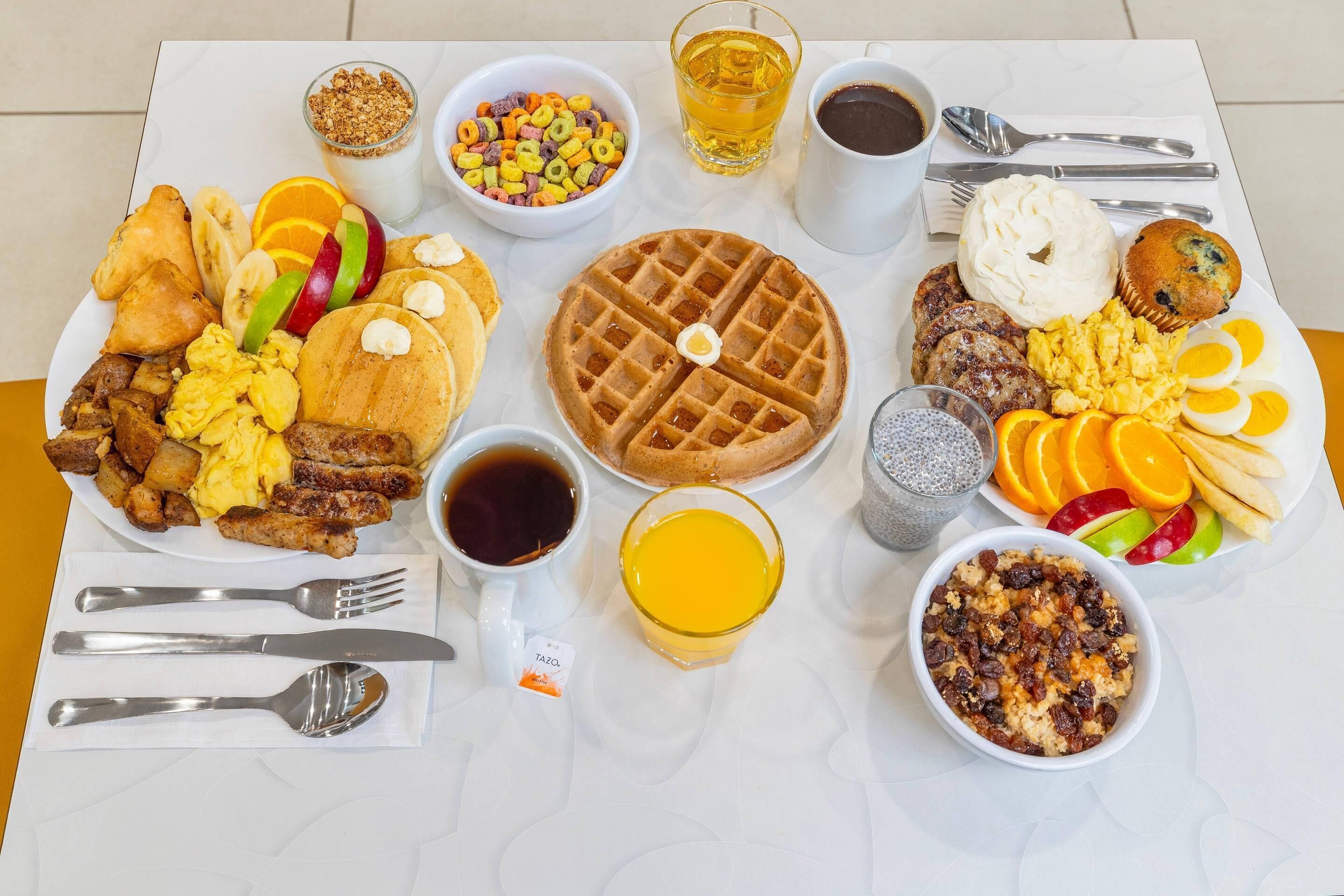 Una mesa con una abundante selección de desayuno, incluyendo waffles, huevos, salchichas y diversas frutas.