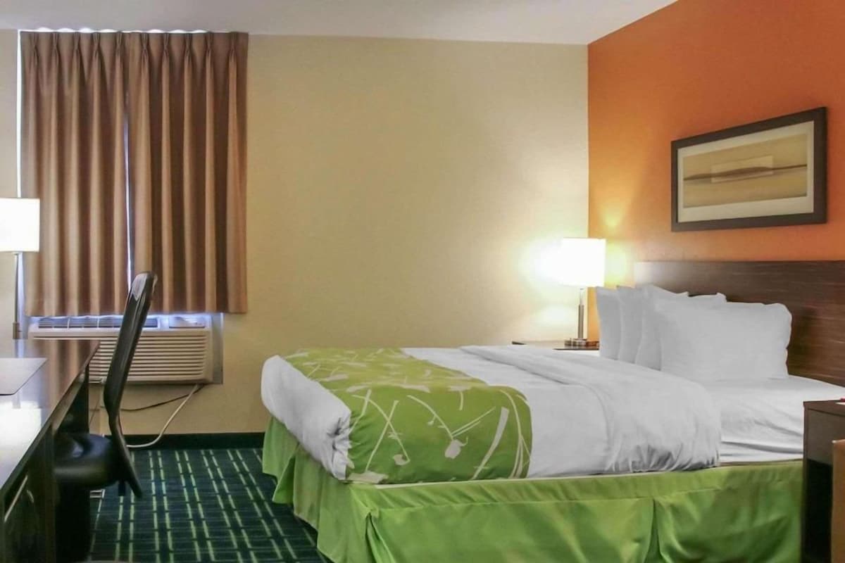 Habitación de hotel con cama, escritorio, silla, lámpara y ventana con cortinas.