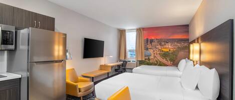 Habitación de hotel con cama, escritorio, silla, televisor, refrigerador y un mural con vista a la ciudad.