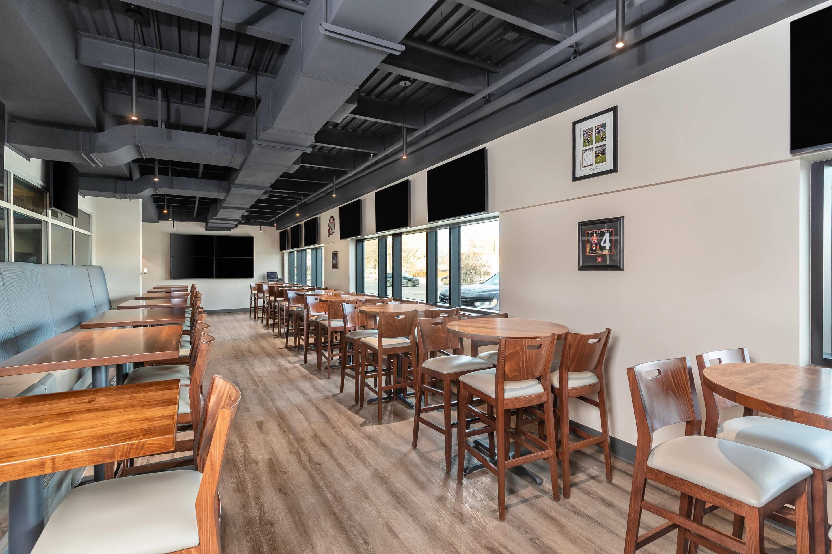 A modern restaurant interior with long wooden tables and chairs, large windows, and exposed ceilings.