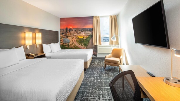 A hotel room with two beds, a desk, a chair, a TV, and a cityscape mural on the wall. - Clarion Pointe (Winnipeg)