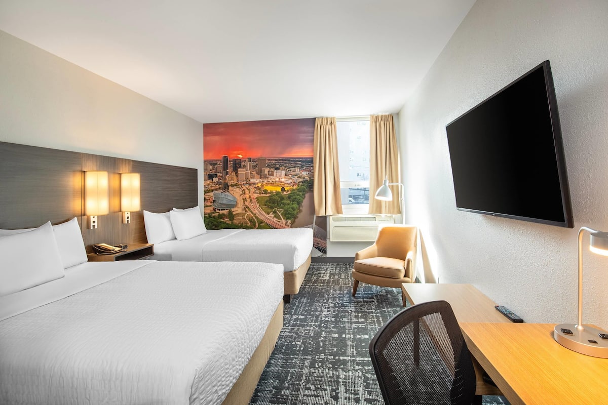 Une chambre d’hôtel avec deux lits, un bureau, une chaise, un téléviseur et une fresque représentant un paysage urbain sur le mur.