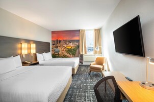 Habitación de hotel con dos camas, un escritorio, una silla, un televisor y un mural con vista a la ciudad en la pared.