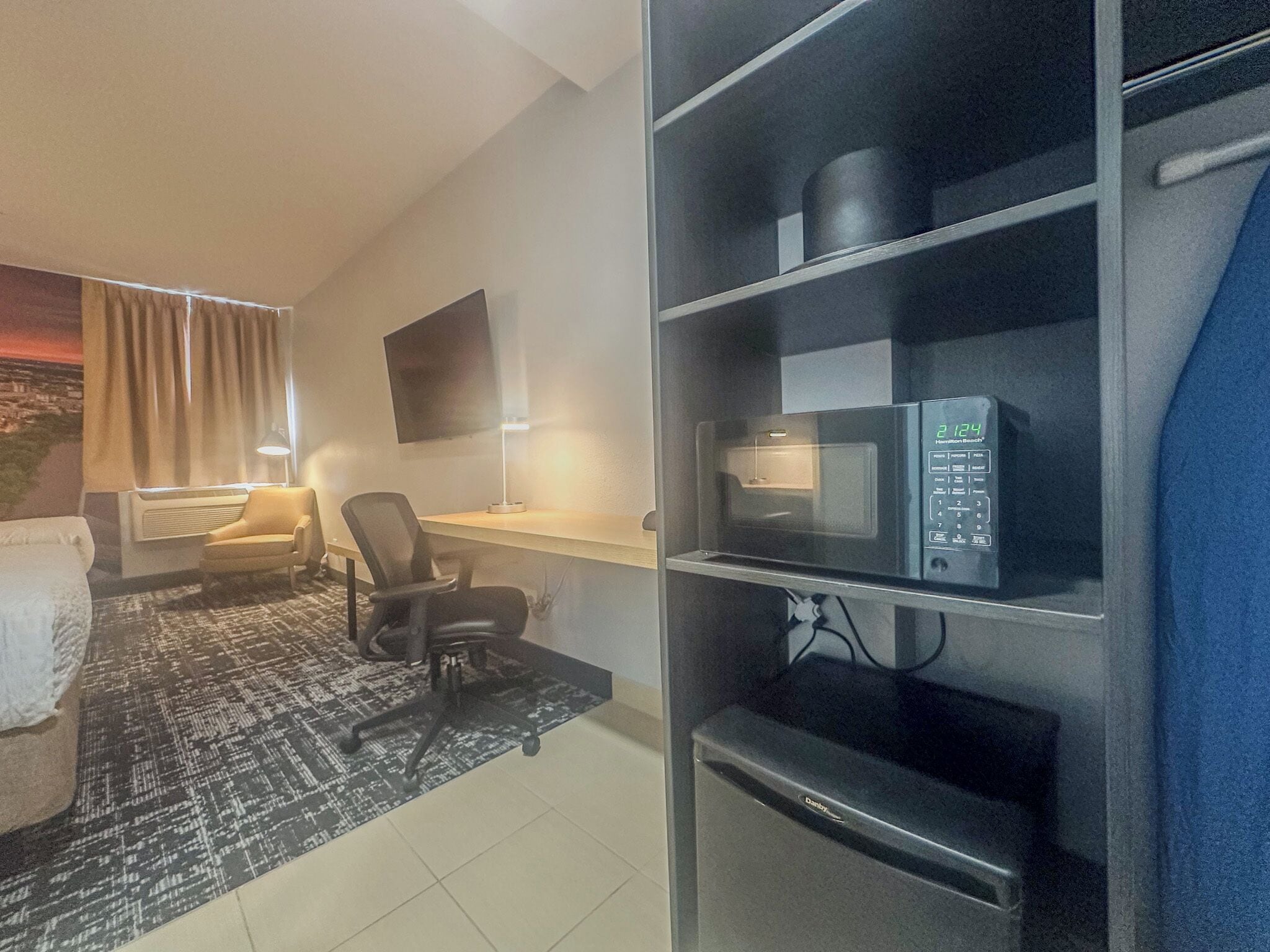 A hotel room with a desk, chair, flat-screen TV, microwave, and a bookshelf.