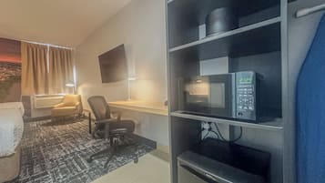 A hotel room with a desk, chair, flat-screen TV, microwave, and a bookshelf.