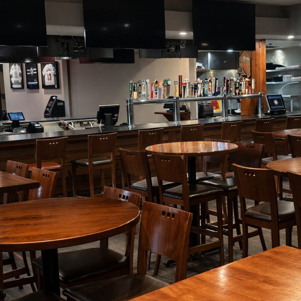 Una zona de bar con mesas y sillas de madera, una barra con caja registradora y una pared decorada con objetos enmarcados.
