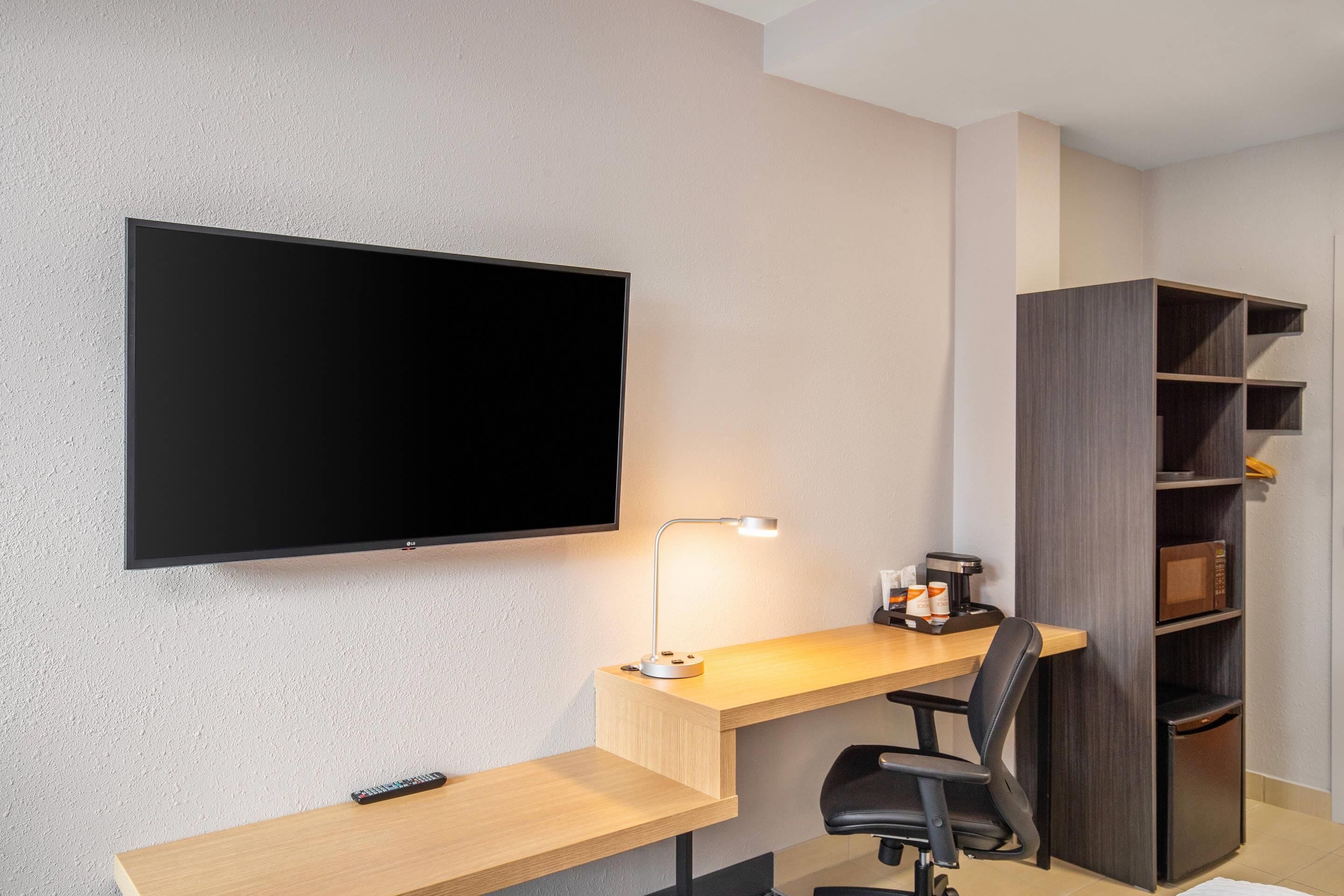 Een moderne hotelkamer met een flatscreen televisie aan de wand, een bureau met lamp, een stoel en een inbouwkast met planken.