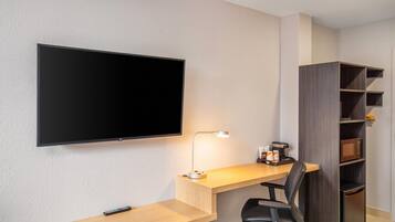 A modern hotel room with a flat-screen TV mounted on the wall, a desk with a lamp, a chair, and a built-in wardrobe with shelves.