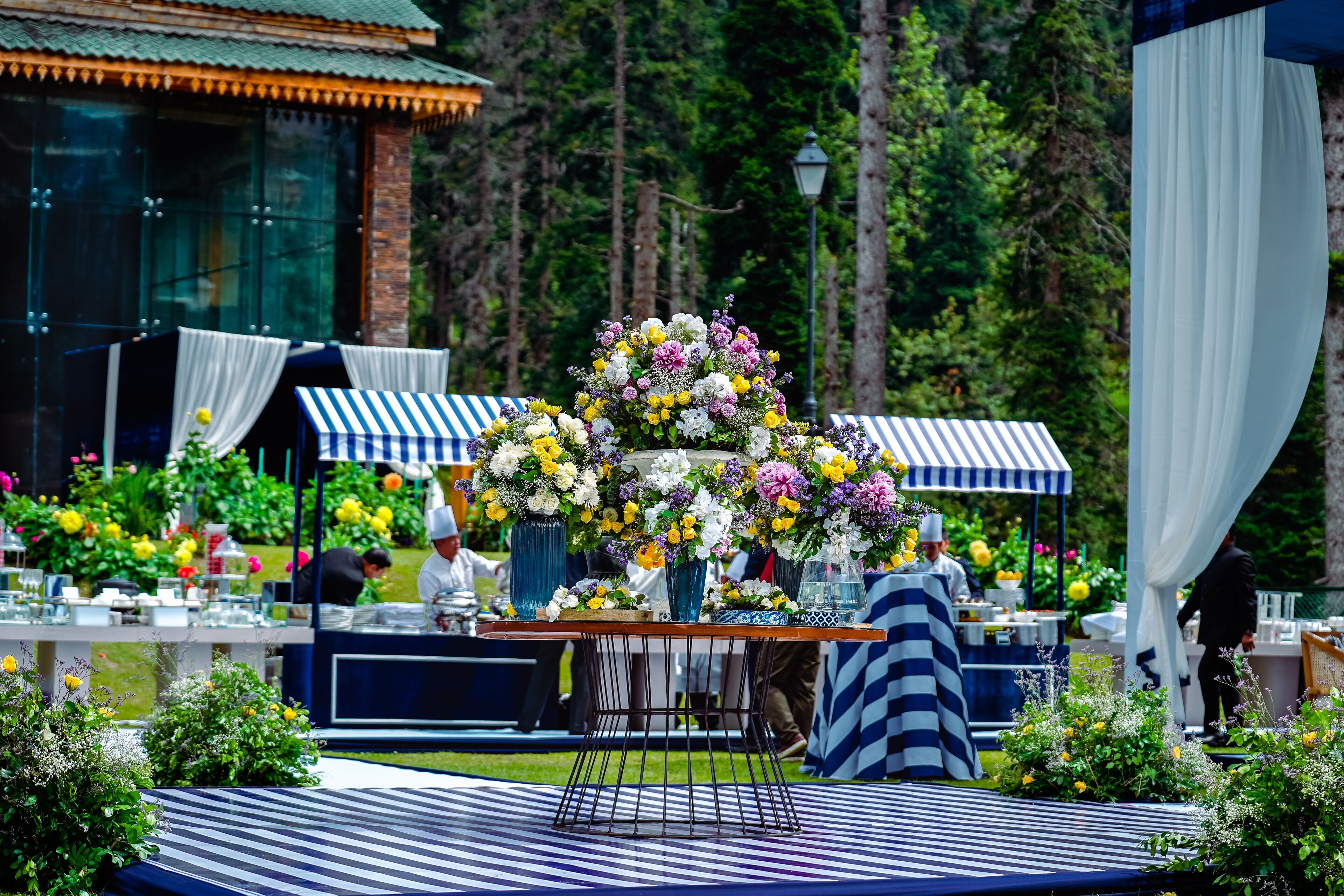 An outdoor event with a central table adorned with floral arrangements, surrounded by striped canopies and tables set up for dining.