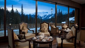 A cozy room with large windows offering a view of snow-covered mountains and a forest. There are elegant chairs and a table set with tea and pastries. - The Khyber Himalayan Resort & Spa (Baramula)