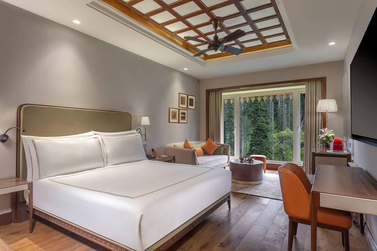 A spacious bedroom with a large bed, a desk, and a chair. There is a window with a view of trees.