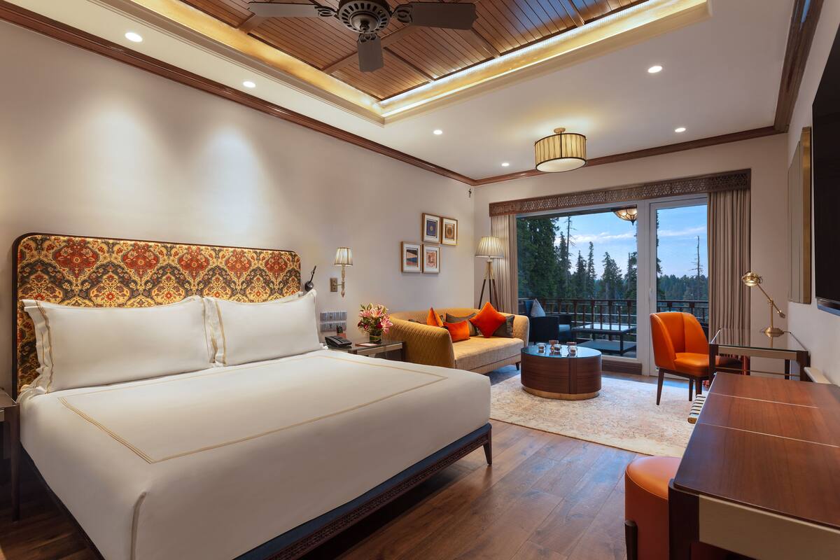 A modern hotel room with a large bed, a sofa, a dining table, and a balcony with a view of trees.