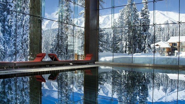 A glass-enclosed indoor pool with a view of snow-covered mountains and a lodge. - The Khyber Himalayan Resort & Spa (Baramula)