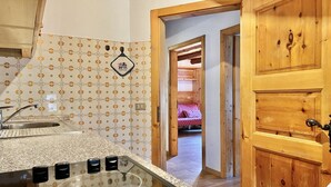 A kitchen with a sink, a stove, and a patterned backsplash. There is a wooden door and a view into a bedroom with a bed.