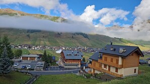 A mountain village with traditional wooden houses, a small parking area, and a view of the surrounding hills and clouds.