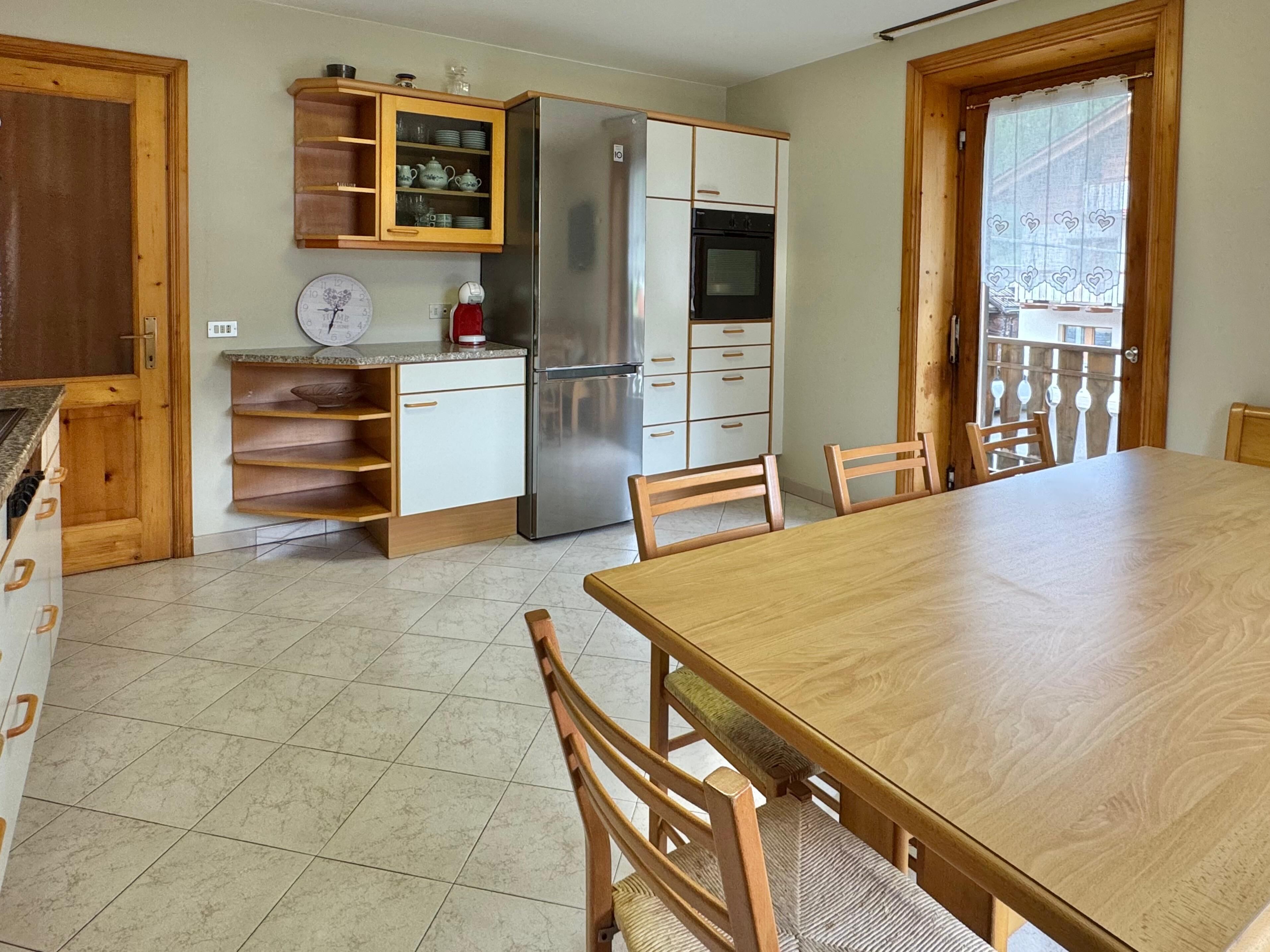 A kitchen with a refrigerator, oven, and wooden cabinets. There is a dining table with chairs and a clock on the wall.