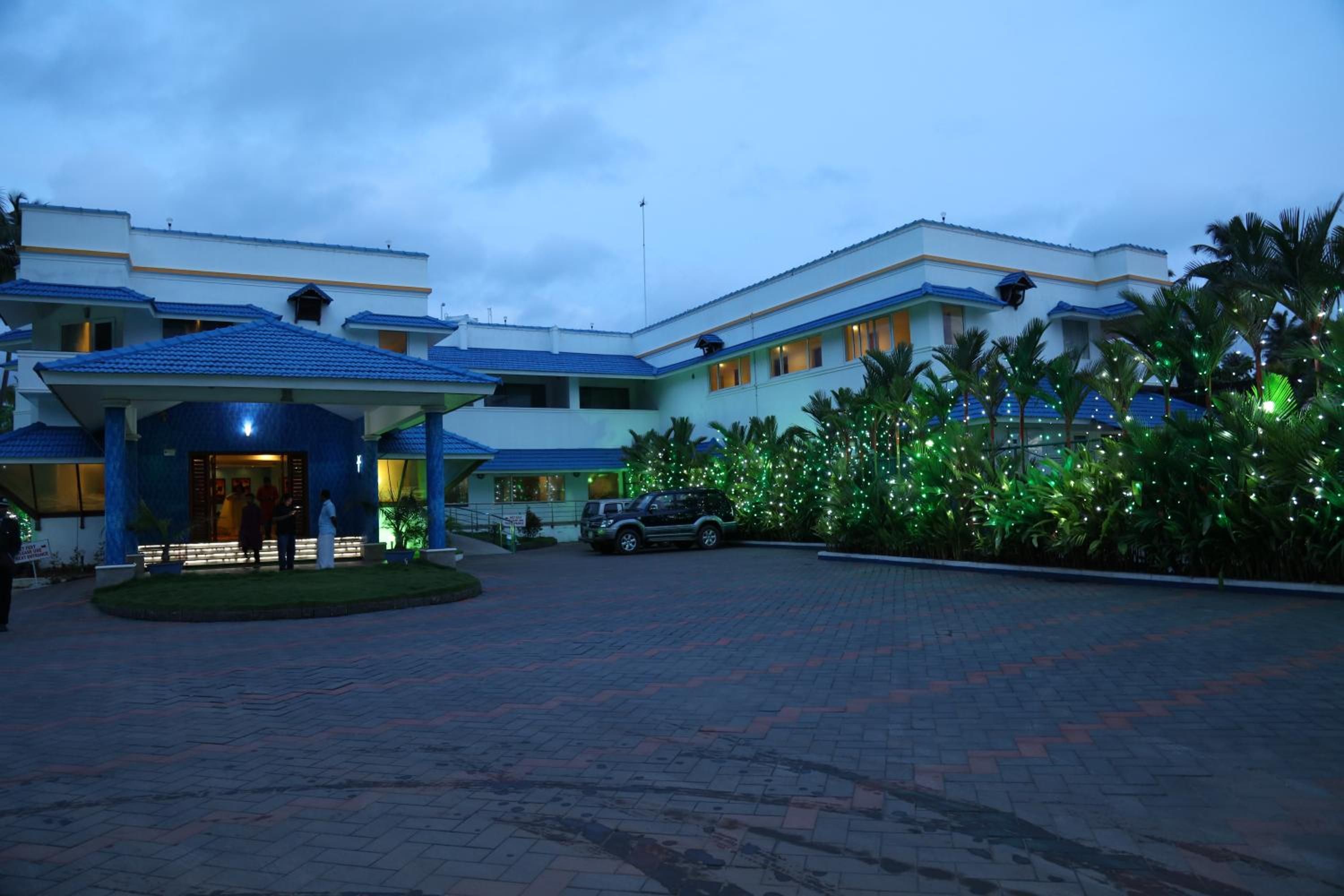 A hotel with a covered entrance, a parking area, and a well-lit garden with decorative lights.