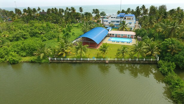 Vista aérea de un resort con piscina, rodeado de vegetación exuberante y palmeras, con vista al mar de fondo.