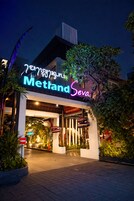 Entrada de hotel com letreiro de neon, acesso coberto e plantas em vasos.