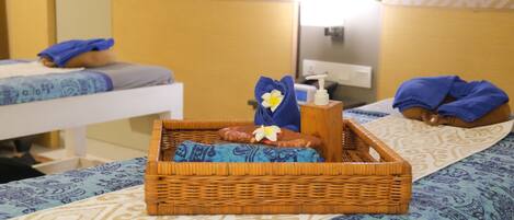 Quarto de hotel com duas camas, cada uma com colcha azul e branca estampada, uma bandeja de vime com uma flor e um frasco de loção.