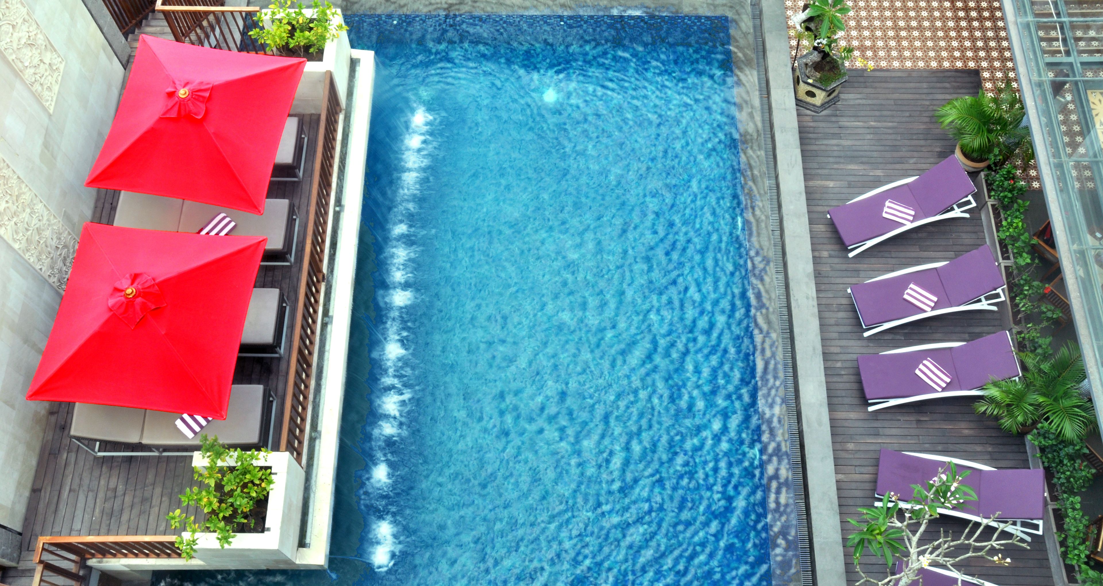 Une piscine rectangulaire à l’eau d’un bleu limpide, entourée d’une terrasse en bois et agrémentée de plantes en pot. Des parasols rouges et des chaises longues violettes sont disposés le long du bord de la piscine.