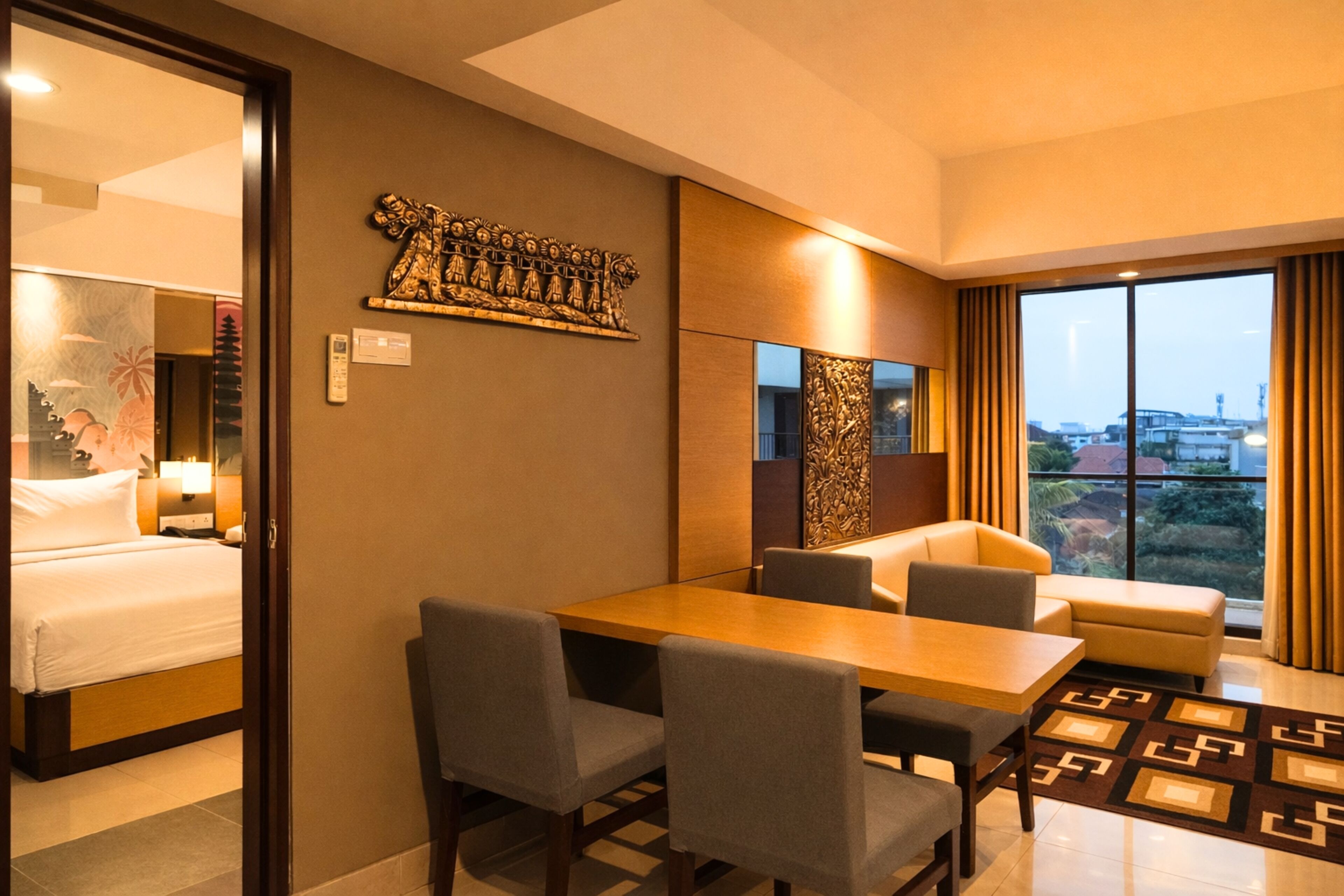 A hotel room with a bed, a dining table with chairs, a sofa, a large window with curtains, and a decorative wall piece.