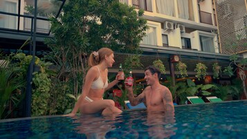 Uma área à beira da piscina com guarda-corpo de vidro, plantas e vista para um edifício com aparelhos de ar condicionado.