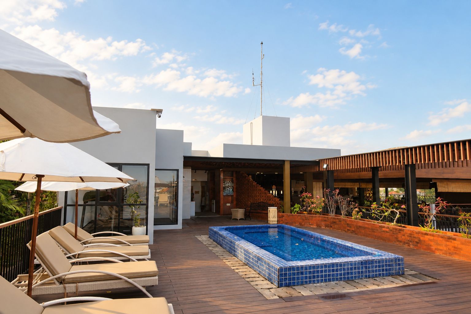 Un área de piscina en la azotea con sillones de descanso y sombrillas, un jacuzzi y un edificio moderno con una escalera que lleva a un nivel superior.