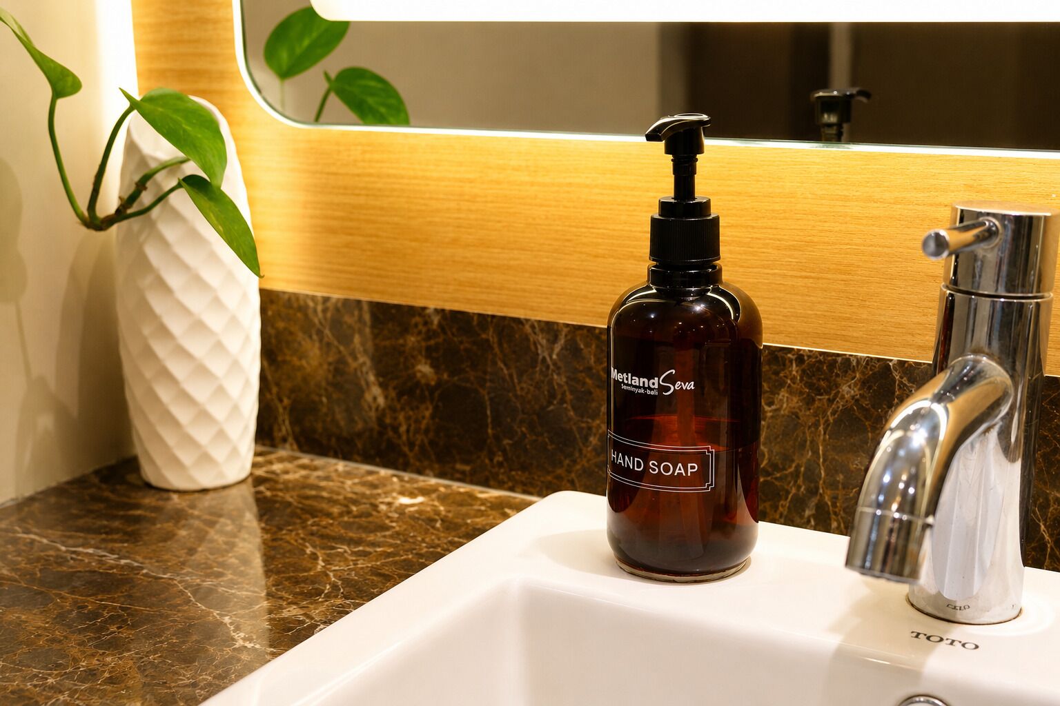 A bathroom with a marble countertop, a white vase with green leaves, a brown hand soap dispenser, and a chrome faucet.