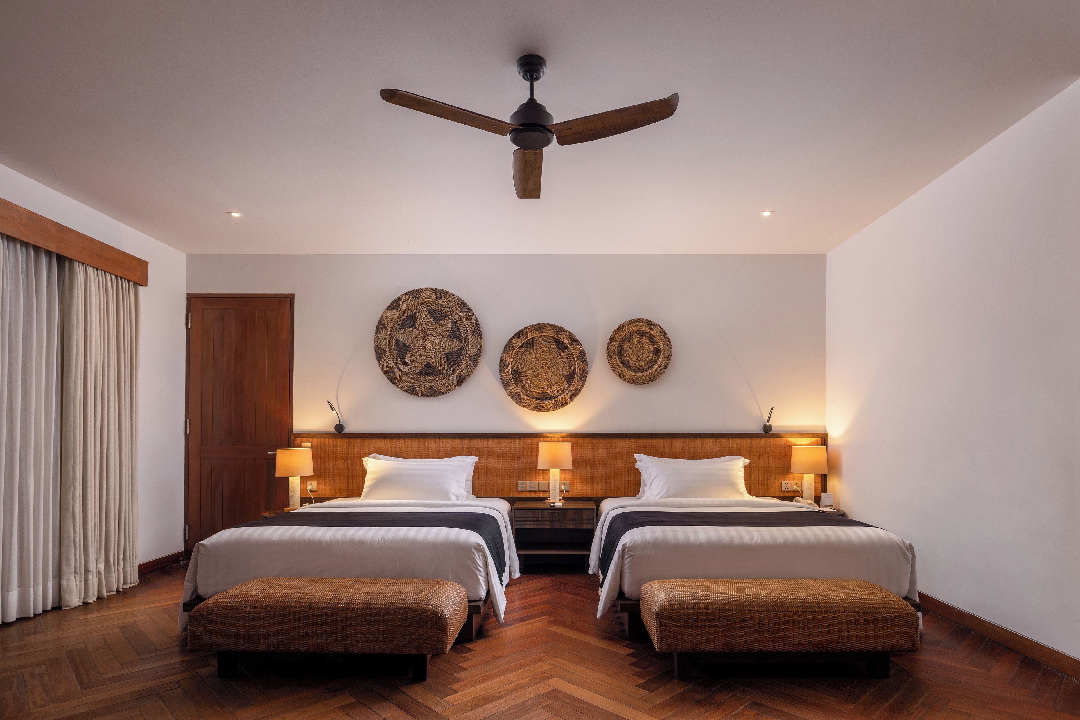 A hotel room with two beds, a ceiling fan, wooden headboard, wall-mounted lamps, and a herringbone wood floor.
