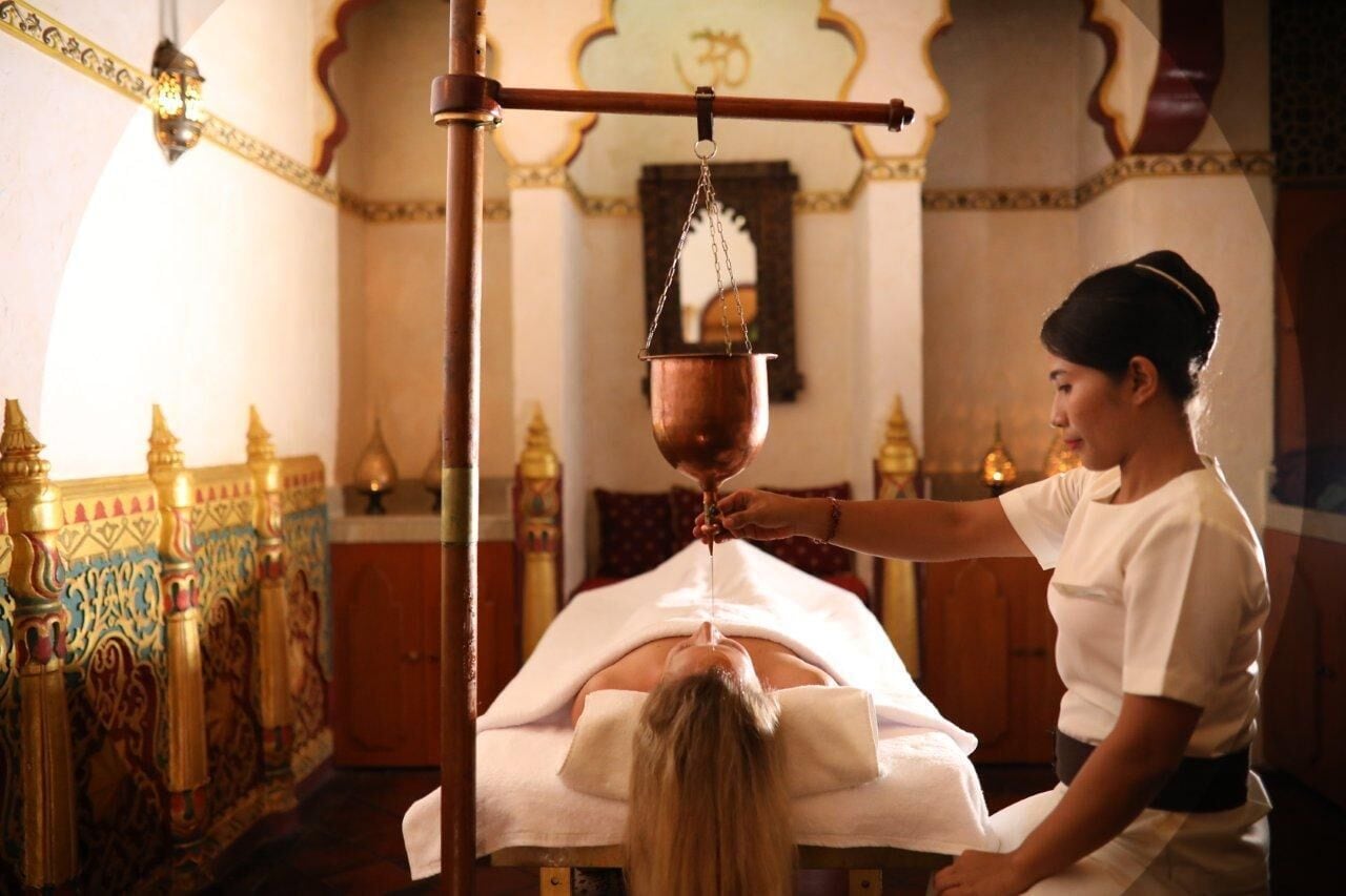 A person receiving a traditional treatment with a copper vessel in a room with ornate columns and a decorative ceiling.