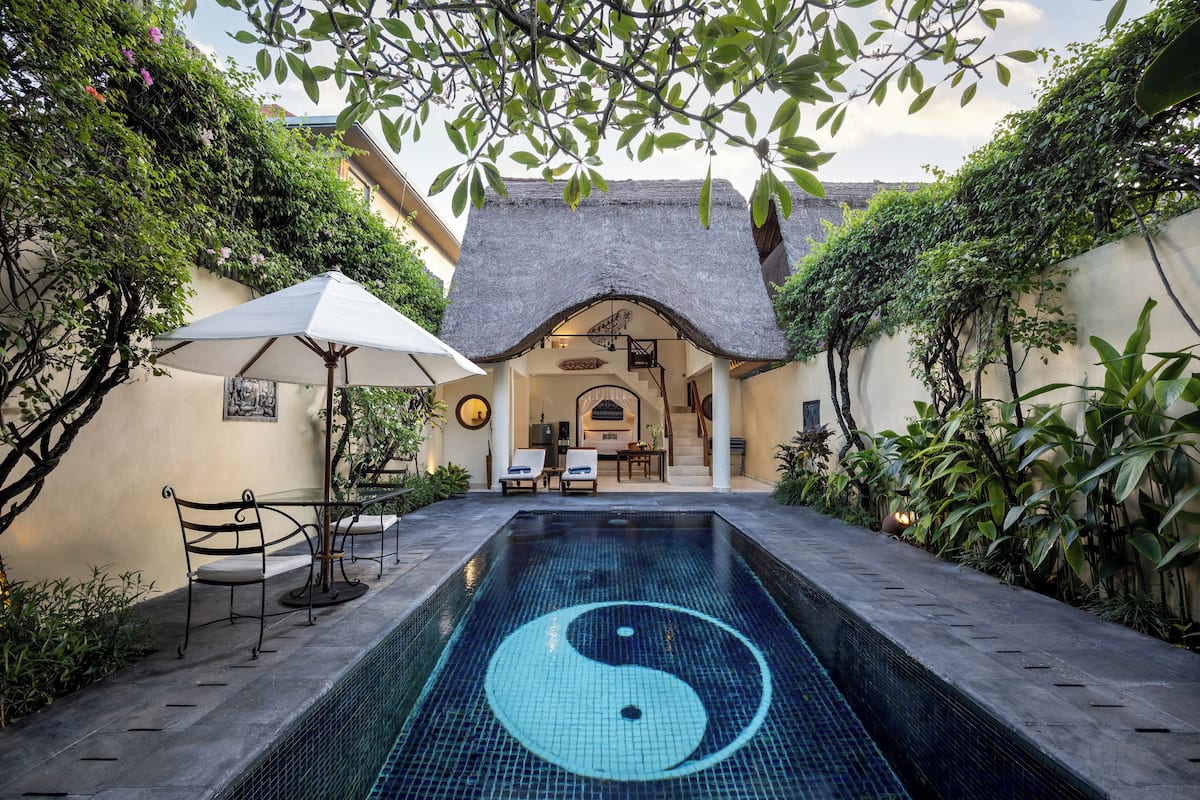 A pool with a yin-yang symbol, surrounded by greenery and a building with a thatched roof.
