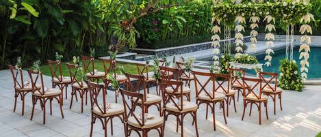 Un espace extérieur aménagé avec des chaises et des tables en bois, entouré d’une végétation luxuriante et agrémenté d’une composition décorative de fleurs et de feuilles.