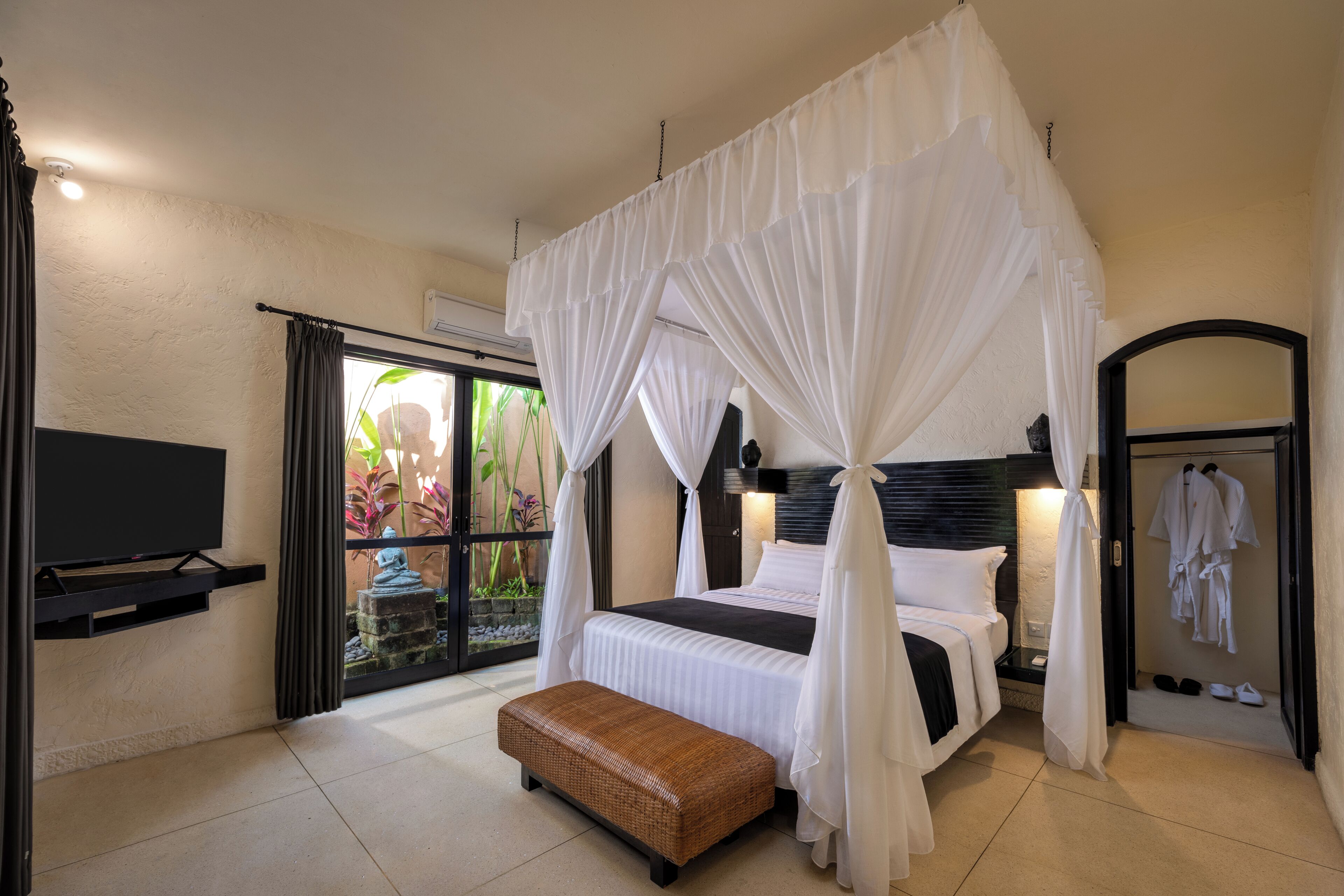A bedroom with a four-poster bed, a flat-screen TV, a bench, and a walk-in closet with robes.