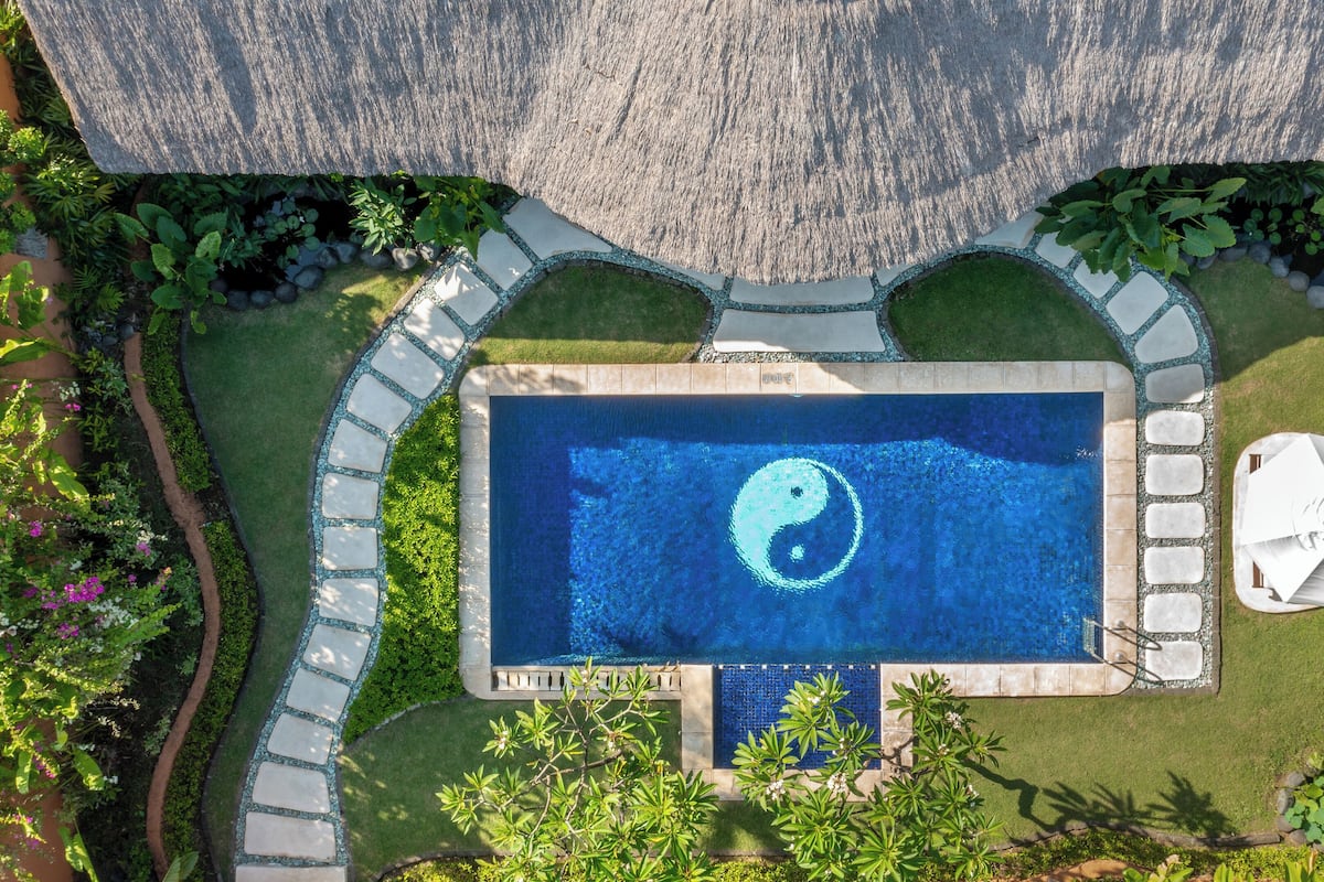 An aerial view of a rectangular swimming pool with a yin-yang symbol in the center, surrounded by a stone border and lush greenery.