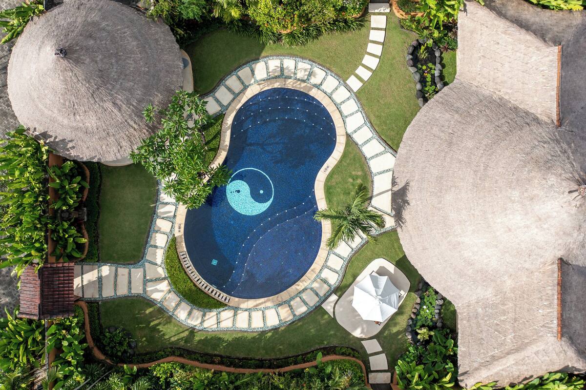 An aerial view of a swimming pool surrounded by a stone pathway, with a thatched roof structure to the left and a palm tree to the right.